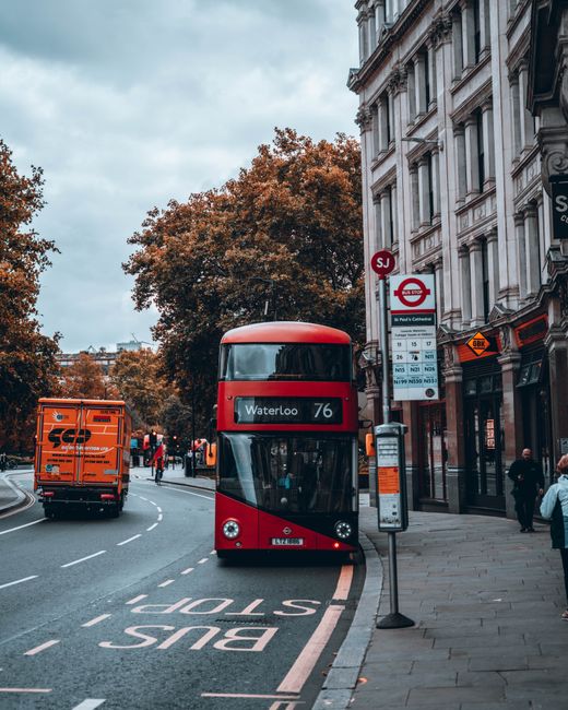 A red double-decker bus displaying 'Waterloo 76' on its digital sign is approaching a bus stop on a city street. The bus stop shelter on the pavement has a timetable attached, and a bus stop sign with the London Transport roundel is visible. To the left, a bright orange delivery van is parked on the road, while several pedestrians walk along the concrete sidewalk adjacent to a historic multi-storey building with ornate facades and large windows. Behind the bus, there are trees with autumn foliage featuring orange and brown leaves, indicating a seasonal setting. The sky above is overcast with grey clouds, and the scene depicts typical urban transport and pedestrian activity. This image relates to house removals and moving services offered by Waterloo Movers, showcasing logistics involved in furniture transport and home relocation processes in Waterloo.