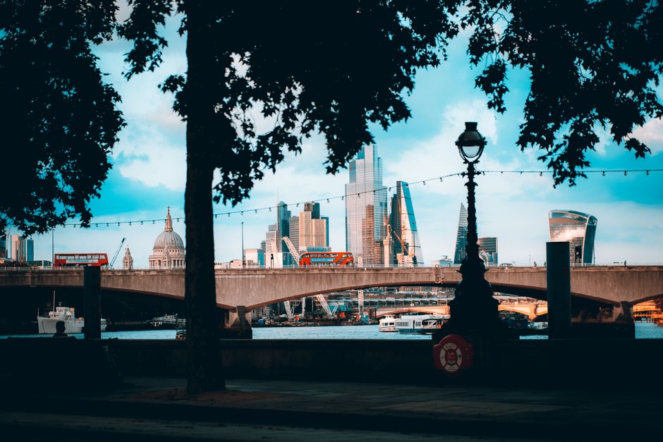 A view of the London skyline with modern skyscrapers, including the Walkie Talkie and Leadenhall Building, seen from across the Thames River during daytime. In the foreground, there are two leafy trees partially framing a paved area with a black lamppost and a life buoy attached to a post. A stone bridge with a low parapet spans across the river, through which several boats and vessels are visible. The scene captures a busy urban environment with a mix of historic and contemporary architecture, under a partly cloudy sky, providing a scenic backdrop for a home relocation or moving process organized by Waterloo Movers.